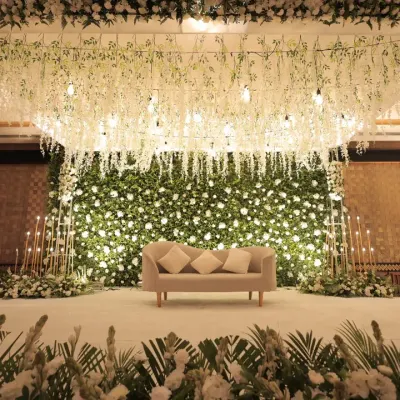 A direct view of our magnificent white floral stage. The design features a dense wall of white roses as a backdrop, with a ceiling of hanging wisteria and delicate lights, creating a breathtaking setting for a wedding or reception.