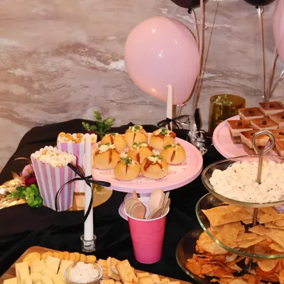 A complete party table setup featuring a mix of sweet and savory options. You can see our pull-apart breads on a pink stand, finger sandwiches, a cheese board, and various dips, all coordinated for a chic black and pink theme.