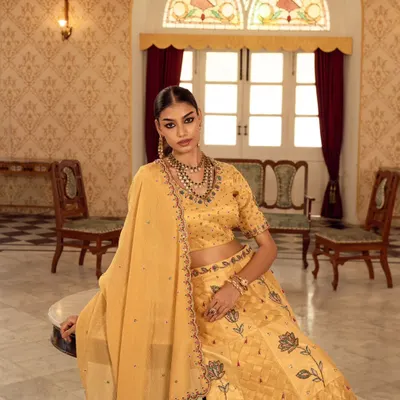 A moment of quiet elegance captured for a luxury ethnic wear campaign. The soft, natural light from the arched window illuminates the detailed embroidery on the yellow lehenga.