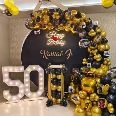 A personalized 50th birthday party featuring a black and gold balloon arch decorated with photos, a neon sign, and large '50' marquee numbers.
