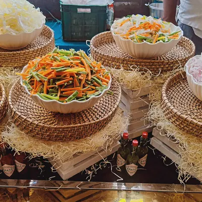 A display of our fresh salad ingredients, featuring bowls of julienned carrots, beans, and shredded cabbage. The rustic presentation with wicker baskets and straw shows our attention to detail in creating an appealing salad bar.