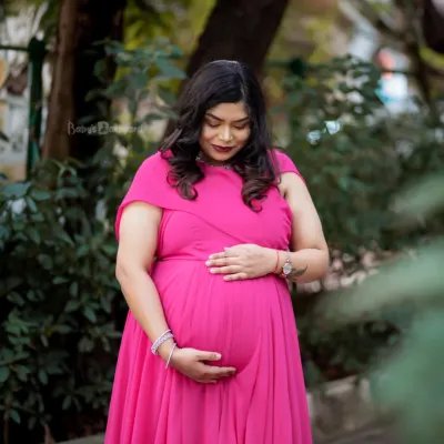 A moment of quiet reflection for the mom-to-be. Dressed in a vibrant pink gown, she gently holds her bump, surrounded by the soft light of an outdoor setting.