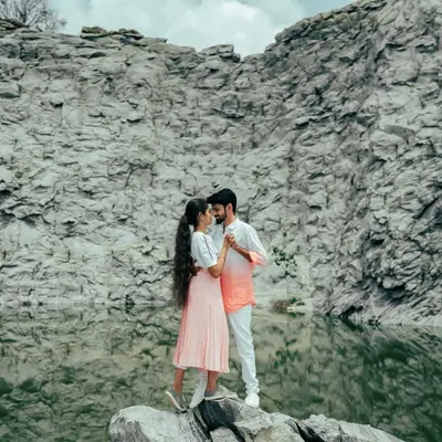 A stunning wide shot of a couple standing on a rock by a tranquil lake, framed by massive rock formations. The scale of the landscape makes this a truly epic pre-wedding photo.