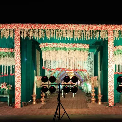 This grand entrance is decorated in a more traditional style, with emerald green drapes and frames covered in red and white flowers. Hanging floral strings, known as 'latkans', add a classic Indian wedding touch to the walkway.
