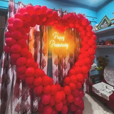 A romantic heart-shaped balloon decoration for a wedding anniversary at home. The frame is made of red balloons with a 'Happy Anniversary' neon sign inside.
