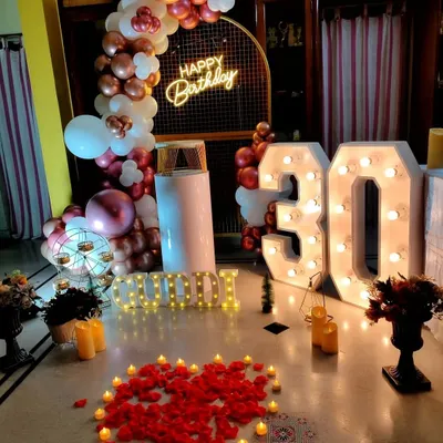 A chic 30th birthday celebration in a modern home. The decor features large light-up marquee numbers, a rose gold balloon arch, and a heart made of petals and candles on the floor, creating a stylish and personal atmosphere.