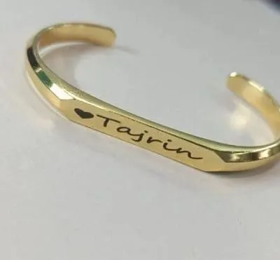 A close-up of a custom name kada in a polished gold finish. The name is engraved in a delicate script and accented with a small heart, creating a simple yet meaningful piece.
