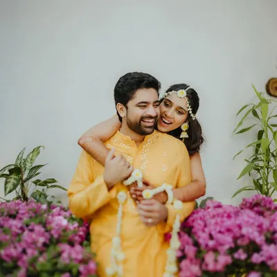 A joyful and candid moment from a Haldi ceremony, with the bride-to-be playfully hugging her partner from behind amidst a backdrop of beautiful purple flowers.