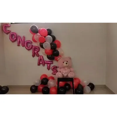 A simple and sweet setup to say 'Congrats'. This corner decoration features pink foil 'Congrats' balloons, a cascade of red, black, and white latex balloons, and a cute pink teddy bear.