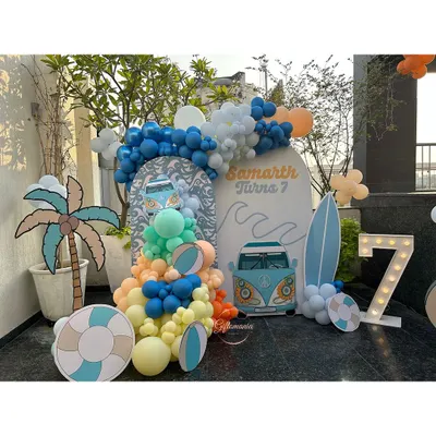 A complete setup for a 7th birthday pool party with a surf and Volkswagen van theme. The decor includes a custom backdrop, a colorful balloon wave, and props like a palm tree, surfboard, and a light up number '7'.