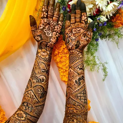 Full-arm bridal henna with a stunning contrast of bold and fine patterns. The design features geometric shapes, paisleys, and floral elements, set against a festive backdrop.