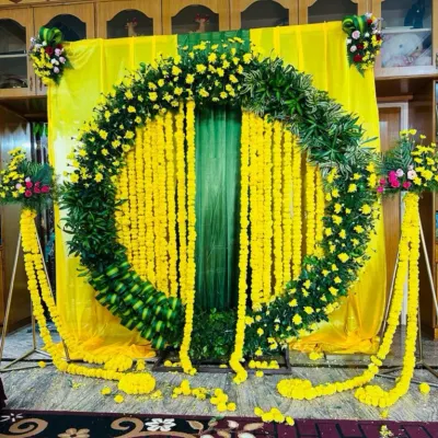 A classic Haldi ceremony backdrop, full of vibrant yellow. This design features a large circular arch made of lush green leaves and yellow flowers, with strings of marigolds hanging in the center against a yellow and green drape.