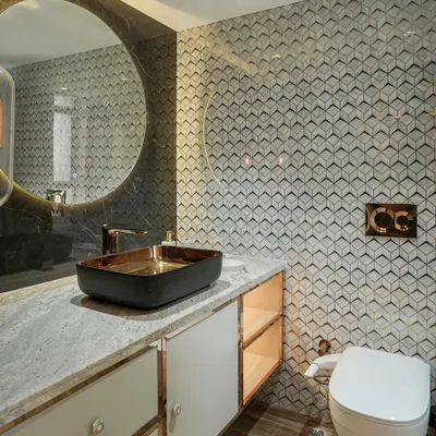 A modern washroom with geometric 3D wall tiles, a black and gold vessel sink, and a large round backlit mirror.
