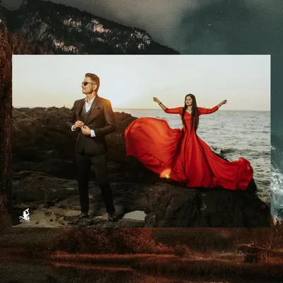 A dramatic pre-wedding portrait on the rocks by the sea. The bride's flowing red dress contrasts beautifully with the dark suit of the groom and the moody sky.