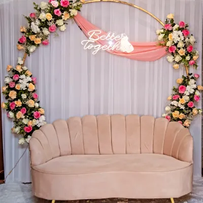 A lovely seating area for the happy couple. This setup features a modern pink velvet sofa in front of a circular golden arch decorated with pink, peach, and white flowers and a "Better Together" neon sign.
