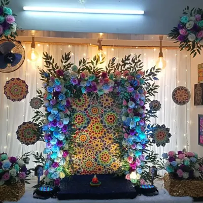 A vibrant home Ganpati decoration with a colorful mandala-style backdrop. The setup is framed by a floral arch and illuminated with blue light for a divine effect.