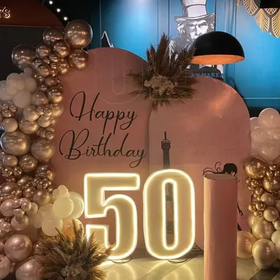 A chic and modern setup for a fabulous 50th birthday. This design features a double U-shaped blush backdrop, a stylish organic balloon arch in champagne gold and white, and dried pampas grass for a touch of boho elegance, all illuminated by a large "50" LED number.