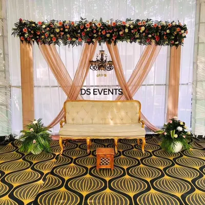 A classic and elegant stage setup. This design features soft drapes, a beautiful floral arrangement overhead, a vintage-style chandelier, and a royal sofa, perfect for a reception or anniversary celebration.