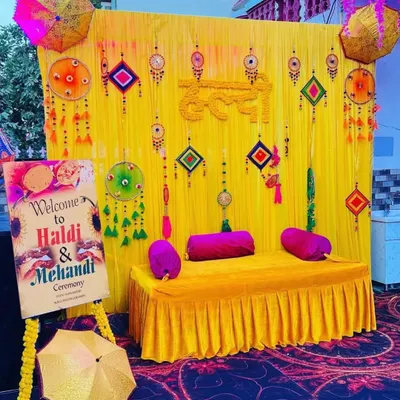 A complete Haldi and Mehendi ceremony setup with a decorated seating bench. The backdrop is a vibrant yellow fabric adorned with traditional hangings, flanked by Rajasthani umbrellas and a custom welcome sign for the event.