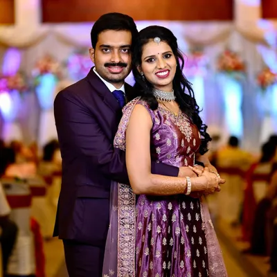 A classic posed portrait of a couple during their wedding reception. The groom is in a sharp suit and the bride wears an elegant purple lehenga, perfectly captured against the stage decor.