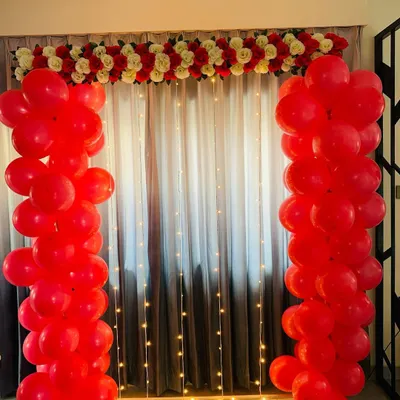 This romantic backdrop features two bold pillars of red balloons, topped with a garland of red and white artificial flowers. The fairy lights on the curtain behind add a soft, warm glow to the setting.