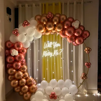 A beautiful indoor proposal setup. This features a large circular ring decorated with a cascade of metallic rose gold and white balloons, with a 'Will You Marry Me?' neon sign in the center.
