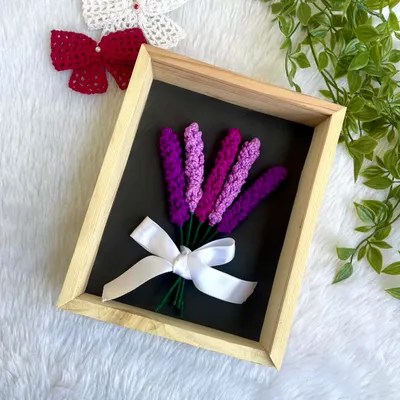 A beautiful arrangement of crochet lavender sprigs, tied with a white ribbon and set inside a wooden frame. This is a simple yet elegant piece of floral wall decor.