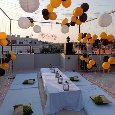 A relaxed and stylish terrace party setup. This decoration features a canopy of fairy lights and paper lanterns, with low seating arrangements and a 'Let's Party' neon sign.
