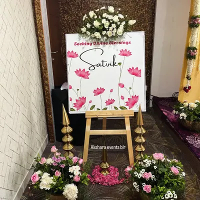 A beautiful welcome sign for a naming ceremony, featuring a lotus theme. The sign is flanked by traditional brass lamps and pots of fresh flowers, creating an auspicious entrance.