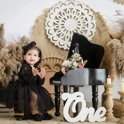 A little star claps her hands with excitement during her boho themed first birthday shoot. The setup includes a miniature piano, pampas grass, and a 'One' sign for a chic and stylish look.