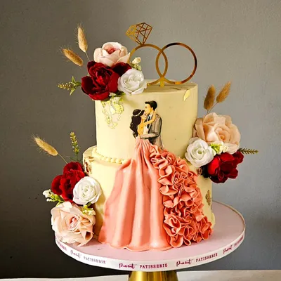 An absolutely stunning engagement cake. This design features a beautiful couple topper with a flowing, ruffled dress, surrounded by handcrafted sugar flowers in shades of red, pink, and white.