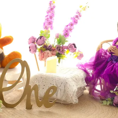 Celebrating her first birthday with a smile and a tea party. This little girl in a beautiful purple dress is surrounded by flowers and her teddy bear companion, marking her special milestone in style.