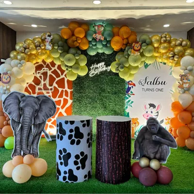 A detailed jungle safari birthday stage. This decoration features a grass wall, animal print pedestals, large cutouts of an elephant and gorilla, and a multi-toned balloon arch for a complete jungle experience.