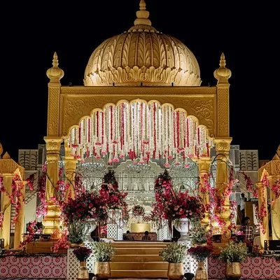 The main wedding mandap, a magnificent golden structure with a central dome and cascading floral garlands. It stands as a beacon of royal elegance against the night sky.