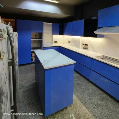 A stunning L-shaped kitchen featuring bold, matte blue Fenix shutters, currently under installation in Vasant Vihar. The design includes a central island and is paired with a white marble-patterned countertop for a striking contrast.