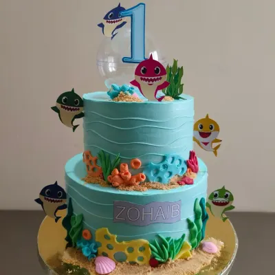 A vibrant two-tier Baby Shark cake, perfect for a first birthday. This underwater scene is created with blue buttercream waves, edible sand, and all the favorite shark characters.