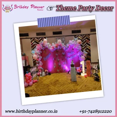 A full view of a pink-themed first birthday party. The decor includes a custom backdrop with balloon garlands, a 'BABY' marquee sign, and a white picket fence prop to create a defined and adorable party space.