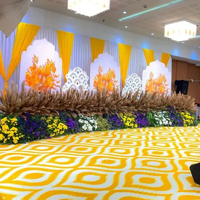 A bright and cheerful stage setup, perfect for a Haldi or Mehndi ceremony. The design uses a vibrant yellow and white color scheme, with a floral base of yellow, purple, and white chrysanthemums mixed with rustic dried pampas grass.