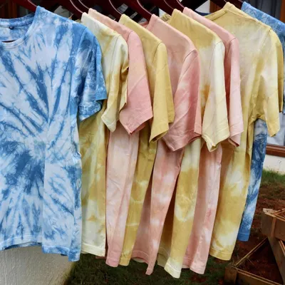 A rainbow of naturally dyed tees hanging in the sun. Each t-shirt is hand-dyed with botanical colors like indigo, madder, and marigold, making every piece unique in its pattern and shade.
