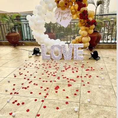 A beautiful rooftop marriage proposal setup. A white and gold balloon arch frames the moment, with 'LOVE' marquee letters and 'Marry Me' sign, all set against a city skyline.