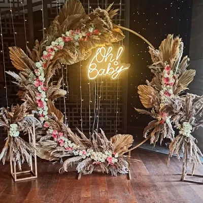 A stunning boho-themed baby shower backdrop with a circular frame, dried pampas grass, and an 'Oh! Baby' neon sign.