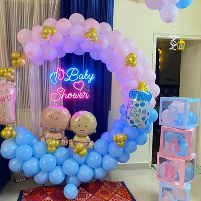 A charming baby shower decoration designed in a cradle shape using pink and blue balloons. The setup includes a 'Baby Shower' neon sign, baby-themed foil balloons, and transparent 'BABY' blocks.