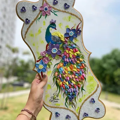 A full view of a 20-inch peacock wall art piece, held to showcase its impressive size and the cascade of multi-colored sculpted feathers. The textured white border adds an elegant finish.