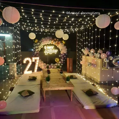 This terrace was converted into a stylish party lounge for a 27th birthday. The setup includes a fairy light canopy, low seating, a balloon ring with a neon sign, and large marquee numbers.