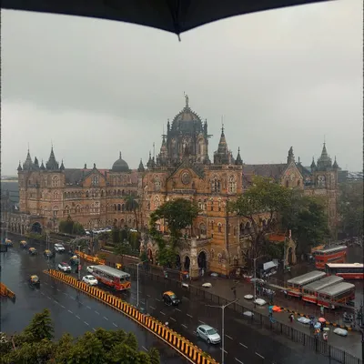 A classic monsoon view of the Chhatrapati Shivaji Maharaj Terminus, framed by the top of an umbrella. This perspective makes the viewer feel like they are right there, witnessing the grandeur of the architecture through the rain.