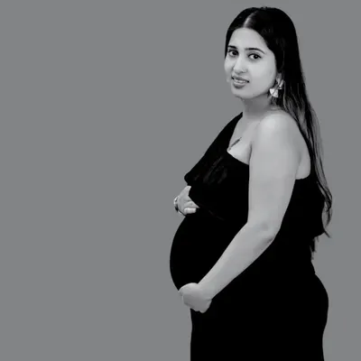 A classic black and white solo portrait. The simple, elegant pose and one-shoulder dress create a timeless silhouette, focusing all attention on the mother's gentle expression and her beautiful baby bump.