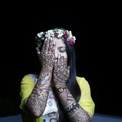 A classic bridal mehendi pose, showcasing the intricate henna designs on both hands. The dark background makes the detailed artwork and the bride's pose stand out.