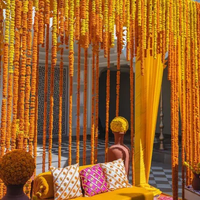 A classic Haldi seating area, defined by a dense curtain of fresh marigold strings. The vibrant yellow sofa and patterned cushions complete this traditional and picture-perfect photo corner.