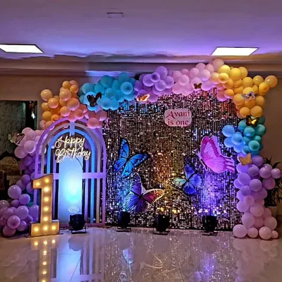 A stunning butterfly theme for Avani's first birthday. This setup features a shimmering sequin backdrop, a custom pastel balloon garland, large butterfly props, a 'Happy Birthday' neon sign, and a '1' marquee letter.