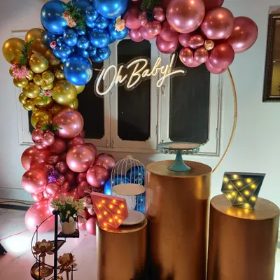 A glamorous 'Oh Baby' shower setup with a circular ring, a cascade of chrome balloons in gold, blue, and pink, and elegant copper-toned plinths.